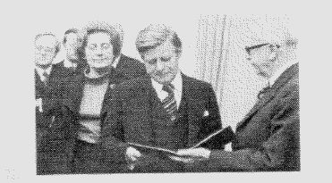 Historisches Schwarz-Weiss-Foto: ein aelterer Herr uebergibt einem Mann ein Dokument, Personen im Hintergrund