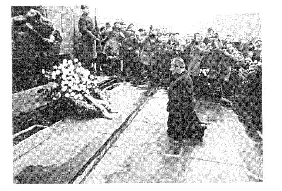 Schwarz-Weiss-Foto: ein Mann kniet vor einem Mahnmal mit Kranz, umgeben von Zuschauern